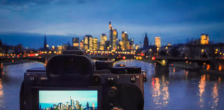 Best Budget Mirrorless Cameras in 2025: Value Testing Guide A full frame mirrorless camera being used on a tripod to capture a long exposure image of Frankfurt's cityscape, with the reflections in the river. The device screen shows the settings to be used - a 13 second exposure at F16, with an ISO of 50.