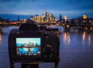 Best Budget Mirrorless Cameras in 2025: Value Testing Guide A full frame mirrorless camera being used on a tripod to capture a long exposure image of Frankfurt's cityscape, with the reflections in the river. The device screen shows the settings to be used - a 13 second exposure at F16, with an ISO of 50.